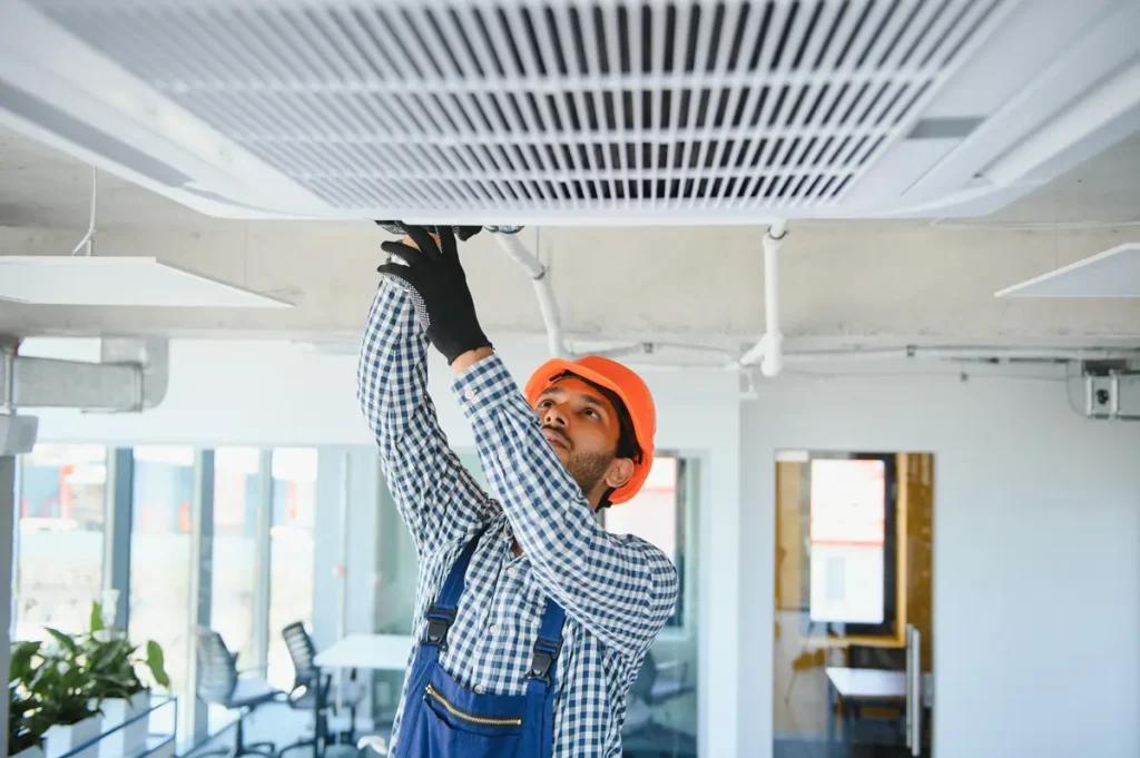 A person in an orange hard hat performing AC repair on a ceiling AC unit.